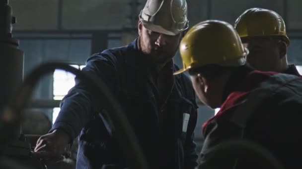 Medium shot van drie trekkerfabrieksarbeiders in coverall kleding en beschermhelmen bespreken werkplan naast moderne machineapparatuur in gieterij werkplaats - Video