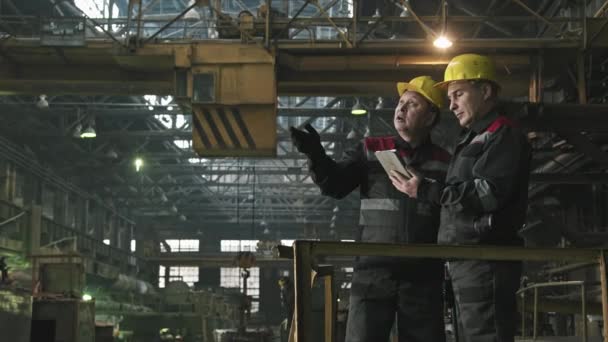 Medium shot of two adult tractor plant workers in uniforms and yellow hard hats standing in foundry shop with digital tablet, looking around and pointing at machine equipment - Footage, Video