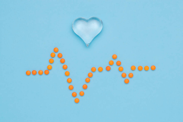 Un dibujo de un cardiograma hecho de pastillas de color naranja y un corazón de vidrio sobre un fondo azul. El concepto de tratamiento de las enfermedades cardíacas. - Foto, imagen