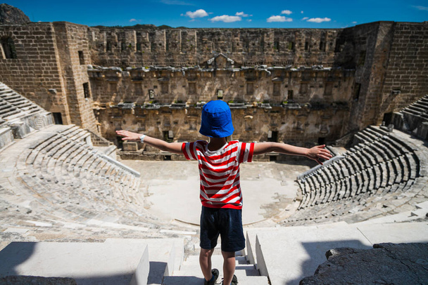 A little boy tourist spread his arms out to the sides against the backdrop of the Roman amphitheater of the ancient city of Aspendos near Antalya, Turkey. Ancient ruined city - Photo, Image