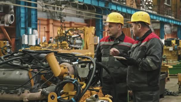 Средний снимок двух работников завода-мужчины в касках и мундирах с использованием цифрового планшета во время осмотра машин в сборочном цехе тракторного завода - Кадры, видео
