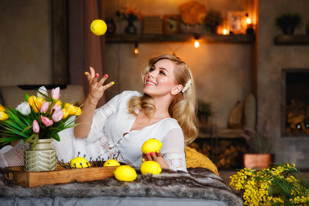 Mooie jonge blonde vrouw liggend op een bed in een landhuis. Vlakbij bloemen en citroenen. Natuurlijke schoonheid. - Foto, afbeelding
