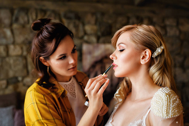 Mooie blonde bruid met make-up en kapsel. Een professionele make-up kunstenaar doet de bruid make-up op de trouwdag. Portret van een jonge vrouw in de ochtend. - Foto, afbeelding