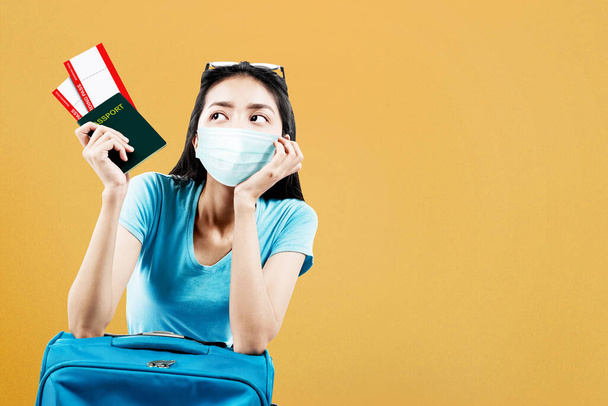 Asian woman in the face mask with a suitcase holding ticket and passport with a colored background. Traveling in the new normal - Фото, изображение