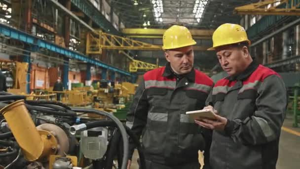 Foto media de dos trabajadores masculinos de la planta en sombreros duros y uniformes conversando mientras inspeccionan equipos de maquinaria en el taller de montaje de una gran planta tractora tomando notas en una tableta digital - Metraje, vídeo