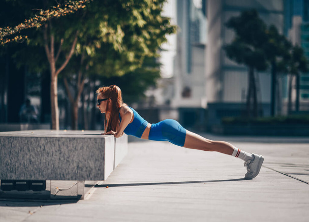 Athlete slim blonde woman fitness trainer in blue sportswear does workout push-ups from bench on city street - Fotoğraf, Görsel