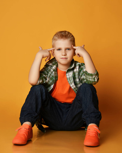 Smart schoolboy teenager in checkered shirt, orange t-shirt and jeans sits on floor holding fingers at head, gesturing - Photo, image