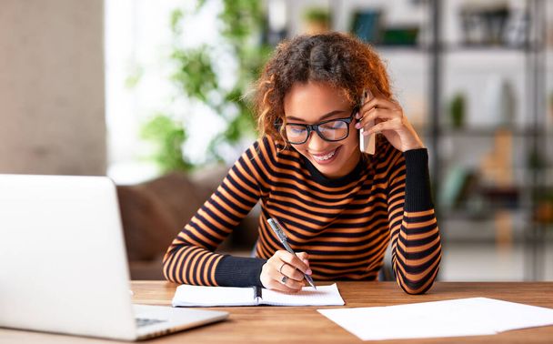 Fröhliche junge afrikanisch-amerikanische Frau, die mit ihrem Kunden auf dem Handy spricht, lächelt und sich Notizen macht, während sie von zu Hause aus am Laptop arbeitet, fröhliche Büroangestellte, die geschäftlich anruft - Foto, Bild