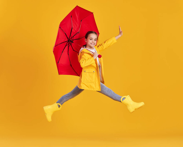 Πλήρης άποψη πλευρά του σώματος του ενθουσιασμένοι κοριτσάκι σε κίτρινο αδιάβροχο και μπότες με κόκκινη ομπρέλα διασκεδάζοντας και πηδώντας πάνω από το έδαφος σε κίτρινο φόντο - Φωτογραφία, εικόνα