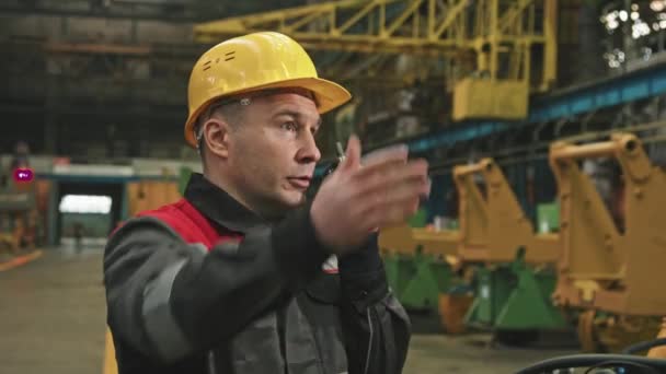 Közepes közel a férfi kaukázusi üzem vezetője gesztikulál, miközben beszél walkie-talkie utasításokat ad az alkalmazottainak dolgozik összeszerelő műhely traktor gyár - Felvétel, videó