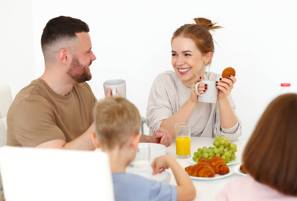 Joven feliz hermosa familia desayunando juntos en casa. Padre, madre y dos lindos niños pequeños comiendo comida saludable en la mañana, hablando y sonriendo mientras están de pie en la cocina moderna - Foto, Imagen