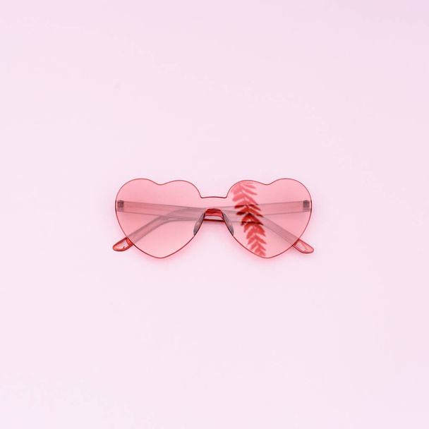 Stylish heart shaped glasses on pink background with copy space. Beautiful trendy red sunglasses and reflection of palm leaf. Fashion summer concept. - Foto, Imagem