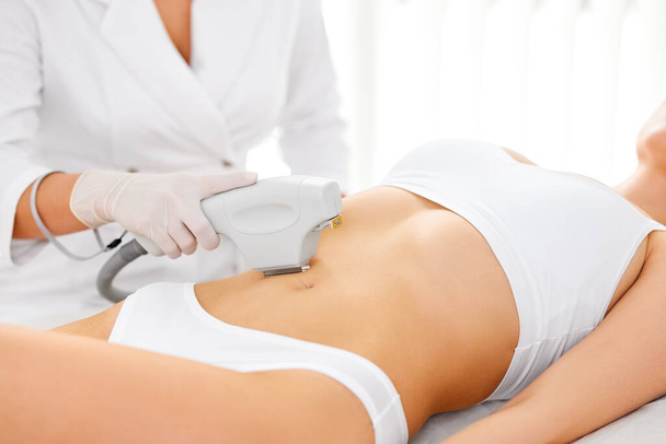 Crop anonymous beautician in gloves using laser hair removal apparatus on bikini zone of female client during beauty laser skin care procedure on stomach  in modern salon - Fotografie, Obrázek