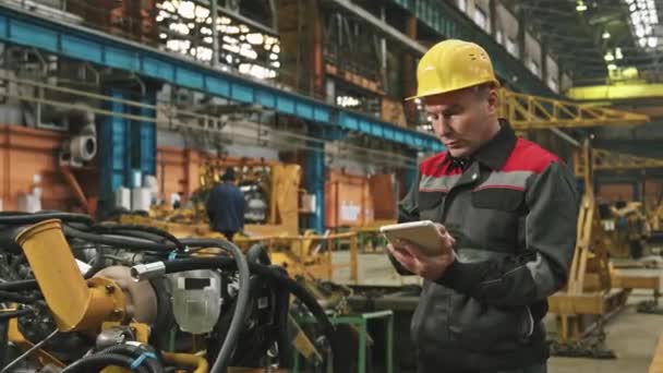 Średnie ujęcie ciężkiego inżyniera zakładu męskiego w twardym kapeluszu i mundurze przy użyciu tabletu cyfrowego podczas kontroli sprzętu maszynowego w warsztacie montażowym dużej fabryki ciągników - Materiał filmowy, wideo