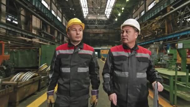 Middelmatige opname van een mannelijke ploegbaas met een harde hoed die een nieuwe arbeider laat zien in de montagewerkplaats van een grote trekkerfabriek die op verschillende machines wijst - Video