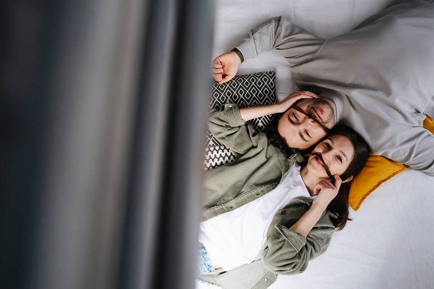Couple chill in a bed. They are lying in an opposite direction, making hair mustache, looking at the camera. Top view. - 写真・画像