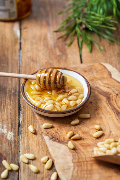 A plate with honey and pine nuts, a honey spoon with transparent honey dripping from it. Light wooden table background, behind a pine branch. - Foto, immagini
