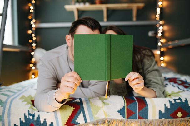 Middle aged couple reading a book in bed, hiding their faces behind the cover. Lying next to each other on a blanket under a garland. - Fotó, kép