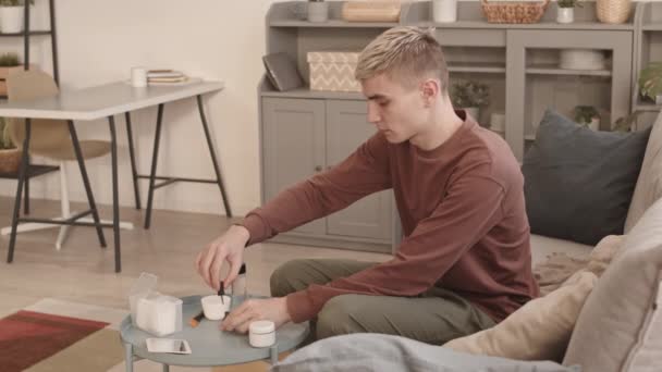 Zamknięcie młodego blondyna białego mężczyzny w ubraniu siedzącego na kanapie w domu i polerującego paznokcie na paznokciach - Materiał filmowy, wideo
