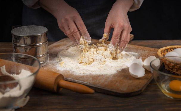 Women's hands, flour and dough. A woman, in an apron prepares dough for homemade baking, a rustic home cozy atmosphere, a dark background with unusual lighting. - Foto, afbeelding