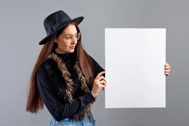 elegant young woman in glasses and hat showing blank board banner, isolated on gray, copy space - Fotoğraf, Görsel