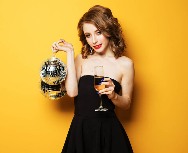 Young blonde woman in evening black dress holding a glass of champagne and disco ball. - Fotografie, Obrázek