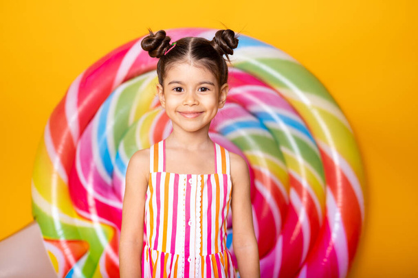 Šťastný vzrušený arabsky dítě dívka v barevné pruhované šaty, které se baví na žlutém pozadí s lízátkem. - Fotografie, Obrázek