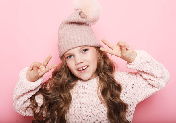 ピンクのセーターと冬の帽子のかわいい女の子が勝利のサインを示す - 写真・画像