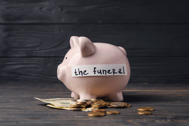 Piggy bank with money and text THE FUNERAL on dark wooden background - Fotó, kép