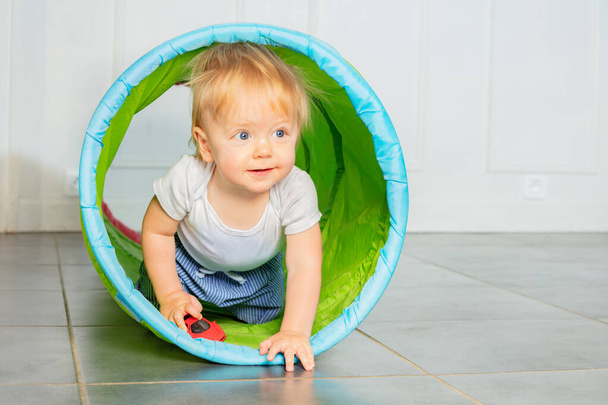 Happy toddler blond boy crawl in the tube with toy car playing in the room - Photo, Image