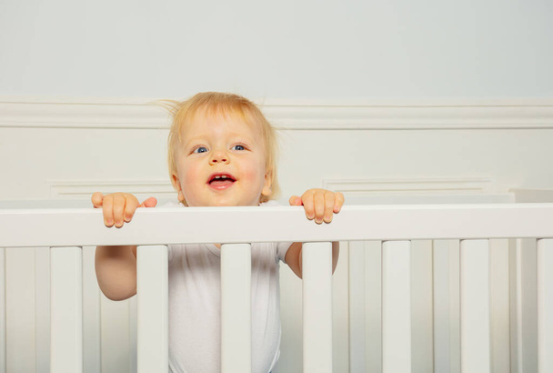 Close face of laughing happy little toddler blond boy stand in the baby crib - Zdjęcie, obraz