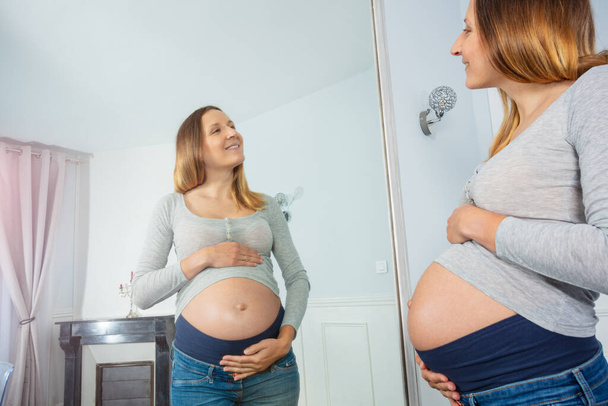 Happy pregnant woman stand before the mirror smiling touching tummy at home - Fotografie, Obrázek