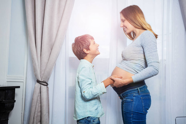 Pregnant woman with son boy holding hands touching tummy near the window - 写真・画像