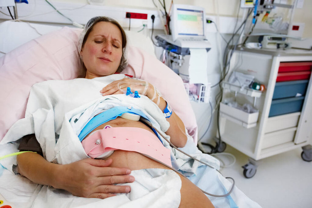Pregnant woman with painful expression and monitors on belly during labor in hospital before giving birth - Foto, Imagem