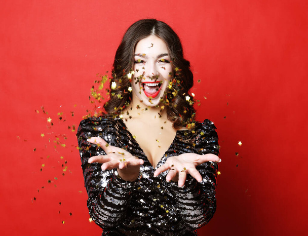 Holiday and party concept: beautiful young woman wearing evening dress standing under confetti rain over red background - Фото, изображение
