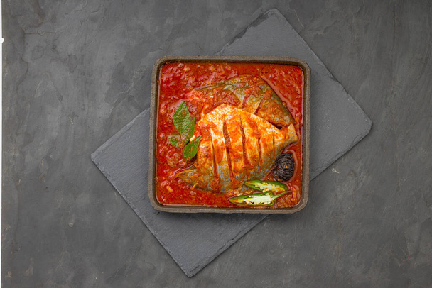  Curry masala pomfret blanco, hecho en estilo indio sur con salsa roja gruesa y adornado con chile verde, hojas de curry y tamarindo malabar dispuestos en un chisporroteo de hierro con fondo de color gris. - Foto, imagen