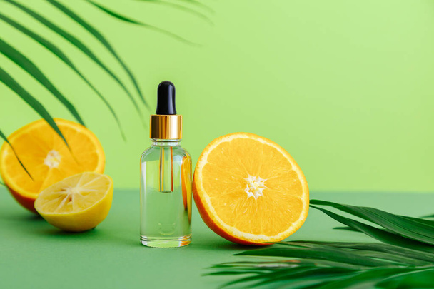 Cosmetic serum Vitamin C in glass bottle with pipette dropper. Orange essential oil with citrus ingredients Vitamin C and palm leaves on color green background. Natural skin care Face Cosmetics. - Fotografie, Obrázek