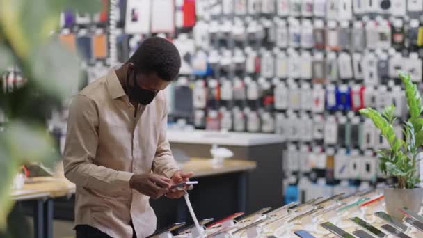 chico afroamericano está de compras durante la pandemia de covid-19 en la tienda de equipos digitales, elegir el teléfono inteligente y pensar en comprar - Imágenes, Vídeo