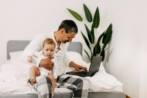 Papa est assis sur le lit, travaille sur un ordinateur portable à la maison avec un bébé sur ses genoux, sourit et regarde l'écran ensemble - Photo, image