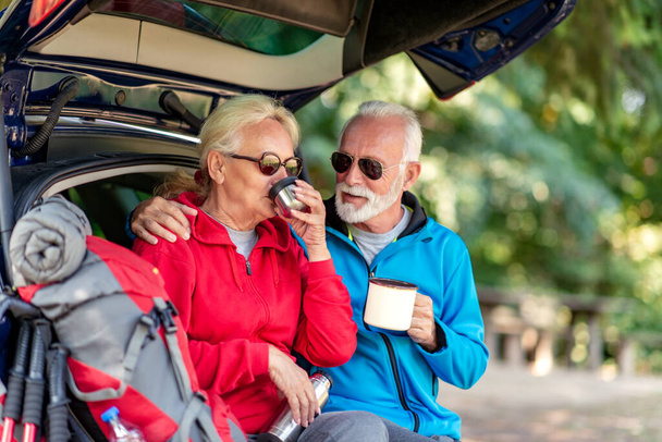 Senior couple sitting in the trunk and drink coffee while resting from hiking. - Photo, Image