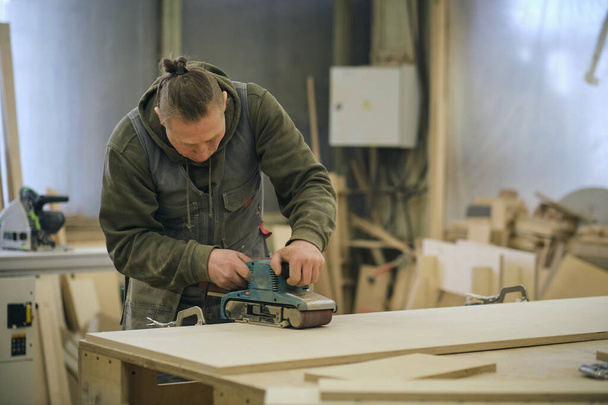 De cerca. Carpintero en taller pule tablero de madera con una lijadora orbital eléctrica. Concepto de carpintería y mobiliario. - Foto, imagen
