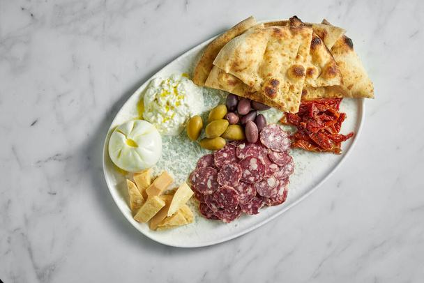 Italian antipasti snacks on white plate on marble background. Focaccia, burrata, olives, sun-dried tomatoes, salami, cheese, parmesan. - Fotografie, Obrázek