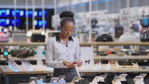 Черная женщина покупатель просматривает выставочные образцы современного мобильного телефона в магазине электроники, дама покупает смартфон в розничном магазине - Кадры, видео