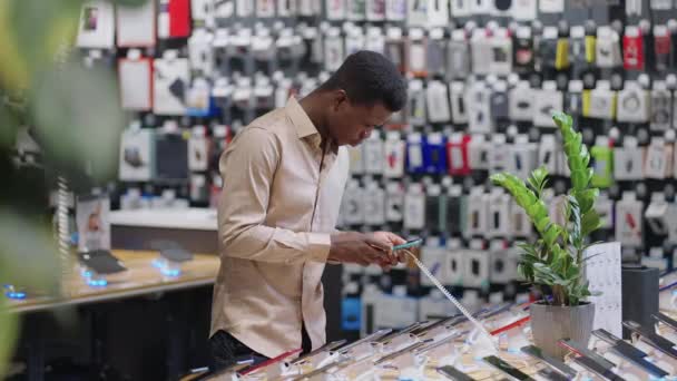 Αφρο-Αμερικανός άνθρωπος δοκιμάζει διαφορετικά μοντέλα smartphones στο κατάστημα ψηφιακού εξοπλισμού, επιλέγοντας συσκευή και σκεπτόμενος την αγορά - Πλάνα, βίντεο