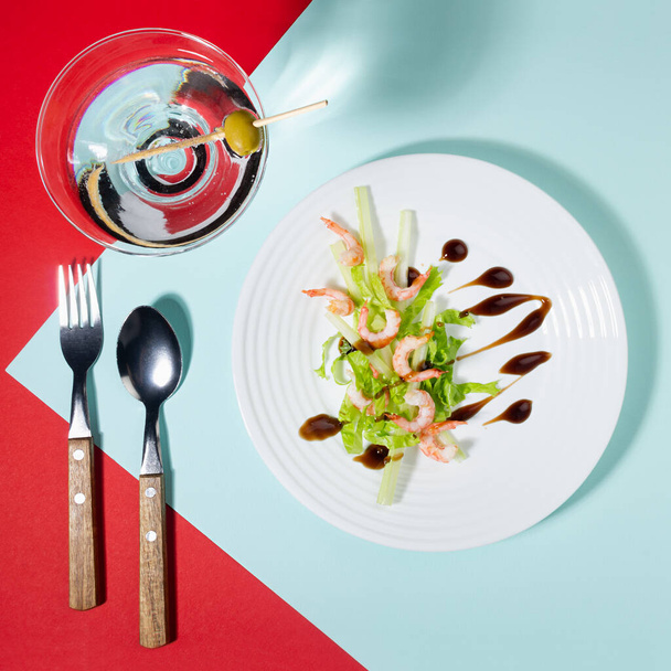 Fresh summer salad of shrimps, greens, celery, teriyaki sauce with cutlery, with martini glass on red and minty color with shadow in sunlight, square. Colorful modern geometric style food background. - Photo, Image