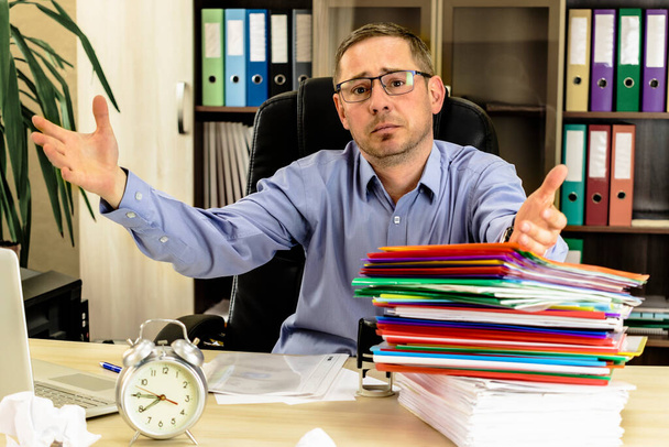 Office worker at his desk in the office with a bunch of folders and crumpled paper. Shrugs his hands not being able to get the job done. Strenuous office work. Financial report. Stress - Photo, Image