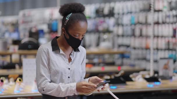ritratto di donna shopper nel negozio di elettronica durante la pandemia di covid-19, donna nera con maschera facciale sta scegliendo smartphone - Filmati, video
