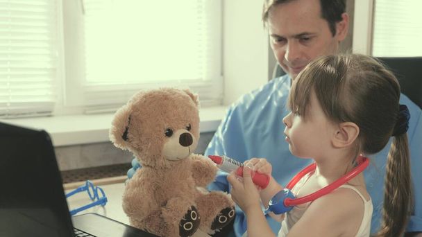 Huolehtiva ammatillinen miespuolinen lastenlääkäri leikkii pienen lapsen kanssa toimistossa. pieni tyttö leikkii nallekarhu lelu kuuntelee häntä stetoskooppi. pikkulapsi potilas istuu lastenlääkärin sylissä. - Valokuva, kuva