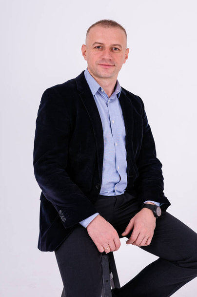 A man in a blue shirt, black trousers and a blue jacket sits on a black stool on a white background - Foto, Bild