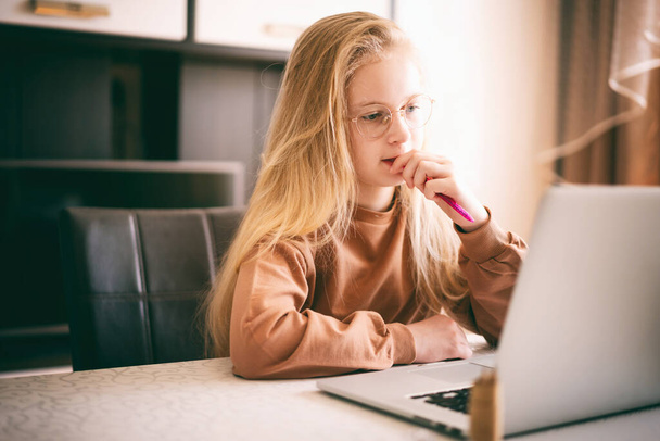 Beautiful 10 years old blond girl in glasses sitting at the desk doing her homework using laptop.. - Valokuva, kuva
